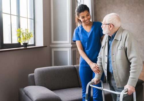 Portrait of an african young nurse helping old elderly disable man grandfather to walk using walker equipment in the bedroom. Senior patient of nursing home moving with walking frame and nurse support