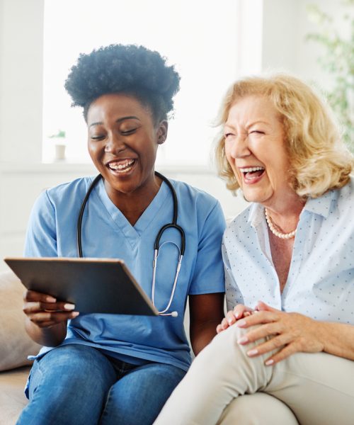 Doctor or nurse caregiver showing a tablet screen to  senior woman and laughing at home or nursing home