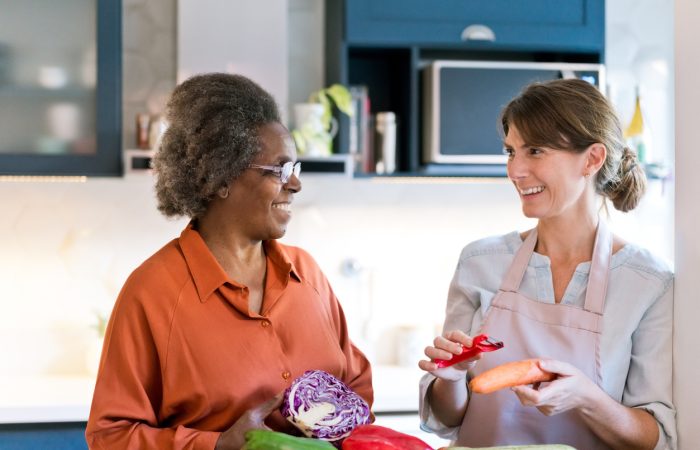 Happy caregiver and senior talking at home. Elderly female and nurse are with vegetables. They are preparing food in kitchen.
