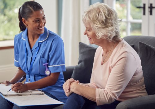 Senior Woman At Home Talking To Female Nurse Or Care Worker In Uniform Making Notes In Folder