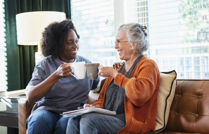 A senior woman in her 80s sitting on the living room couch, having coffee and conversing with a home healthcare aide, an African-American woman in her 30s.