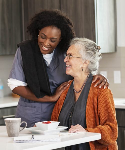 A senior woman in her 80s sitting at home in her kitchen eating a healthy snack. A home caregiver is standing beside her and they are both smiling. The healthcare worker is an African-American woman in her 30s.