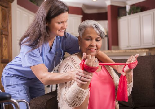 Caring, Latin descent home healthcare nurse conducts physical therapy exercises with African descent senior adult patient at home, assisted living, or nursing home setting.  She helps her with arm strengthening exercises using resistance band.  Kitchen background.  Wheelchair foreground.