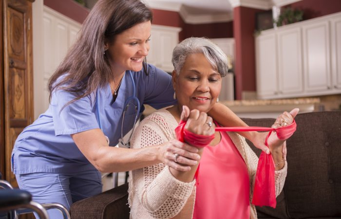 Caring, Latin descent home healthcare nurse conducts physical therapy exercises with African descent senior adult patient at home, assisted living, or nursing home setting.  She helps her with arm strengthening exercises using resistance band.  Kitchen background.  Wheelchair foreground.